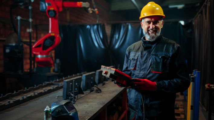 A mature factory worker is standing next to a robotic welder and holding the controlling console in hand.