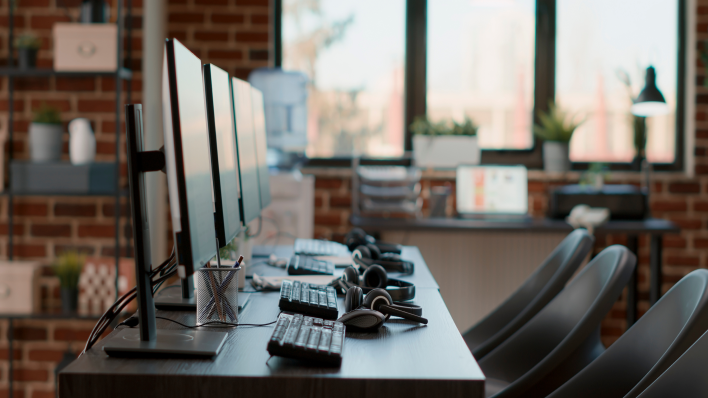 Row of 3 empty workstations with keyboards and headsets lying on desktop. There is a window and brick wall in the background. 