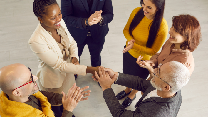 Overhead shot of six people in a circle, congratulating a black woman in a beige suit.