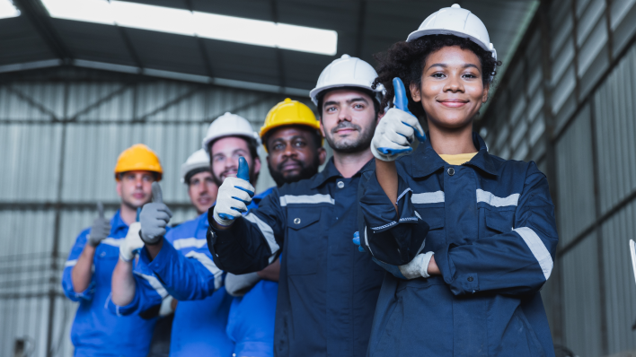 A line of 6 multicultural factory workers in hard hats and work gloves. All are pointed at the camera giving a thumbs up.