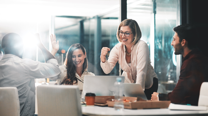 Navigating Talent Shortages 4 people (2 men, 2 women) in modern conference room. Smiling blonde woman is making a fist pumping motion and the woman to her right is about to high five man across the table.