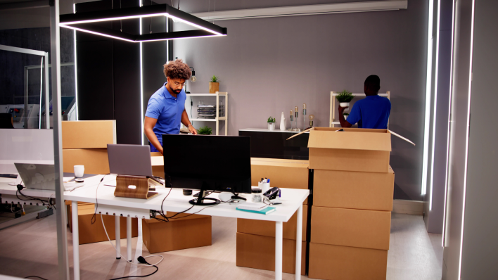 Two male movers in blue shirts packing up an office. Boxes are stacked on the floor and monitors visible on desktop.