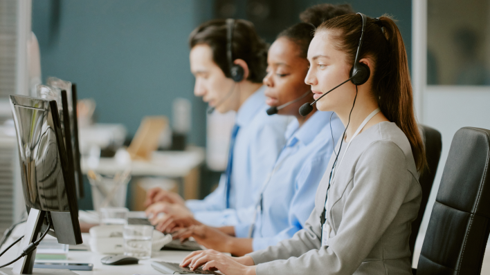 Three call center employees in a row sitting at terminals with headsets on: a white woman, black woman and dark-haired