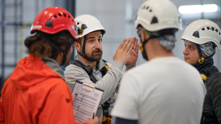 3 people in hard hats gathered around an instructor also wearing a hard hat. He is making a gesture by holding his hands in an upward-pointing position. One of the onlookers holds a clipboard. 